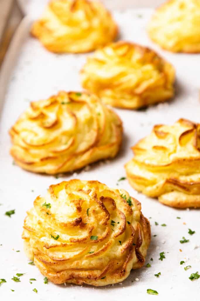 Golden duchess potatoes beautifully arranged on a parchment-lined baking sheet, piped in an elegant spiral shape. These crispy-edged duchess potatoes are garnished with fresh chopped herbs, adding a touch of green to their perfect golden hue.