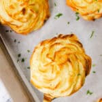 Close-up of freshly baked, golden brown Duchess Potatoes on a baking tray lined with parchment paper. This delightful potato recipe features one lifted on a spatula, showcasing its swirled, piped design and finishing touch of sprinkled chopped herbs.