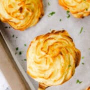 Close-up of freshly baked, golden brown Duchess Potatoes on a baking tray lined with parchment paper. This delightful potato recipe features one lifted on a spatula, showcasing its swirled, piped design and finishing touch of sprinkled chopped herbs.