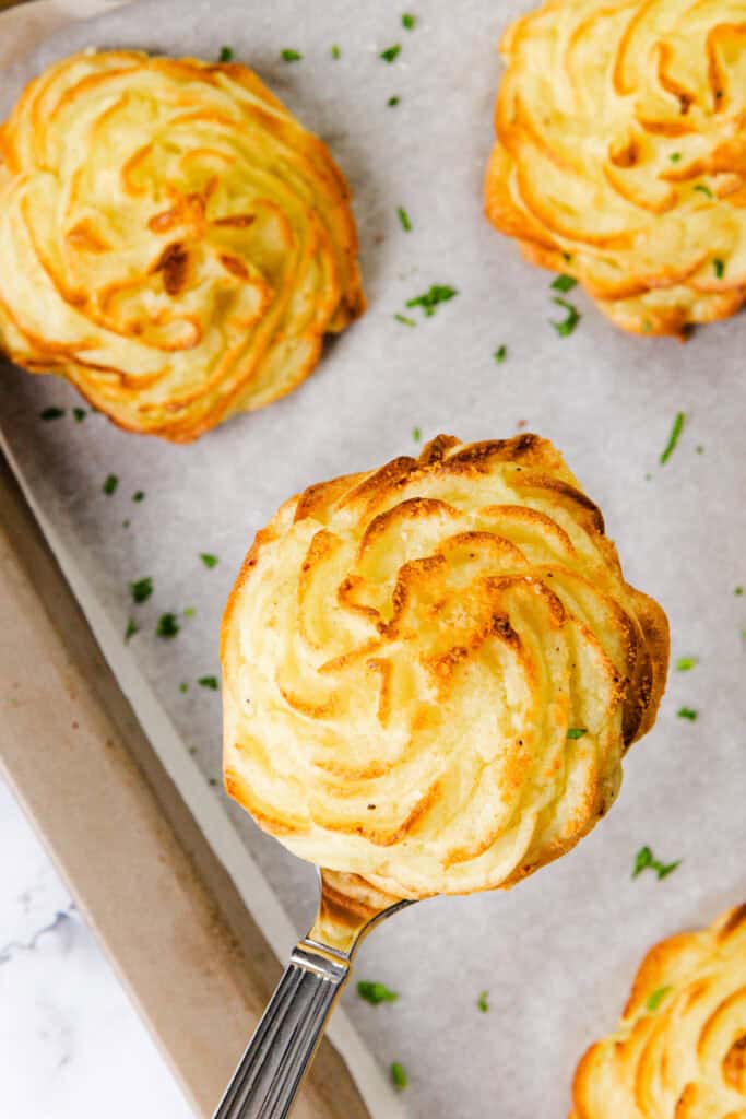Close-up of freshly baked, golden brown Duchess Potatoes on a baking tray lined with parchment paper. This delightful potato recipe features one lifted on a spatula, showcasing its swirled, piped design and finishing touch of sprinkled chopped herbs.