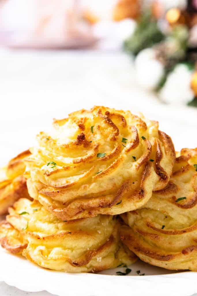 A close-up of golden brown duchess potatoes, intricately piped like a gourmet mashed potatoes recipe, and garnished with chopped herbs on a white plate. The background features a blurred festive setting.