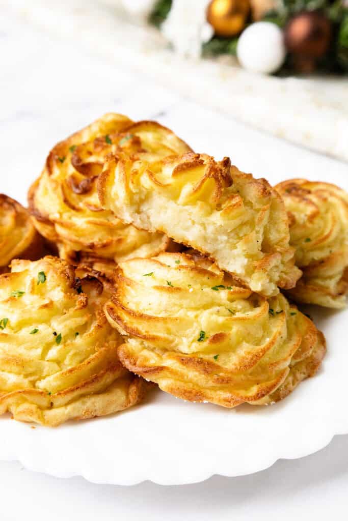 A close-up of golden-brown Duchess Potatoes showcases their elegance. These mashed wonders are piped into swirled shapes, baked to crispy perfection, and garnished with herbs. One is halved, revealing the fluffy interior—a true feast for the eyes and palate.