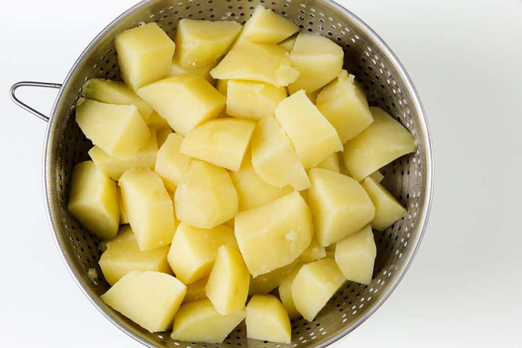 A metal colander filled with peeled, boiled potato chunks sits on a white surface, ready to be transformed into a gourmet side dish.