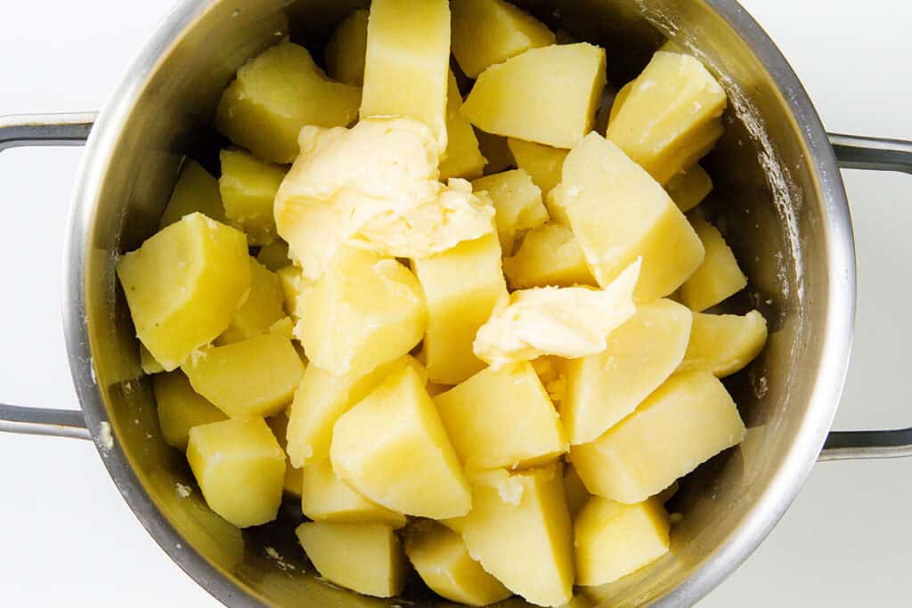 A pot filled with boiled potato chunks topped with a dollop of butter, ready for mashing into a gourmet side dish. The pale yellow potatoes and slightly melting butter set the stage for a delightful Duchess Potatoes recipe.