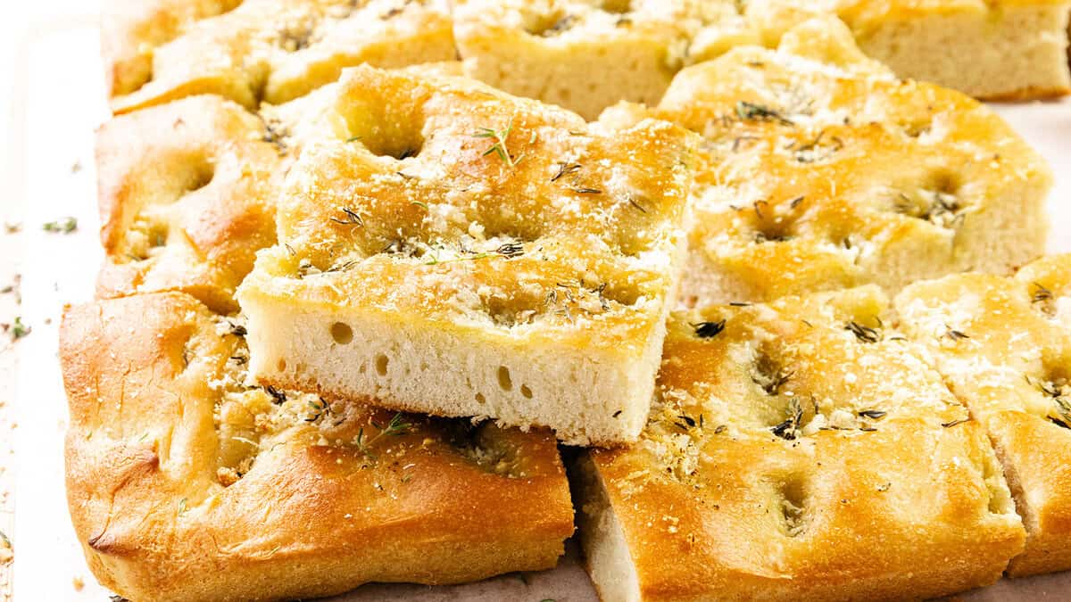 Golden, fluffy squares of focaccia bread are stacked on top of each other, topped with herbs and grated cheese—perfect for adding to your collection of Thanksgiving recipes, with their soft interior and lightly crisp, golden-brown crust.