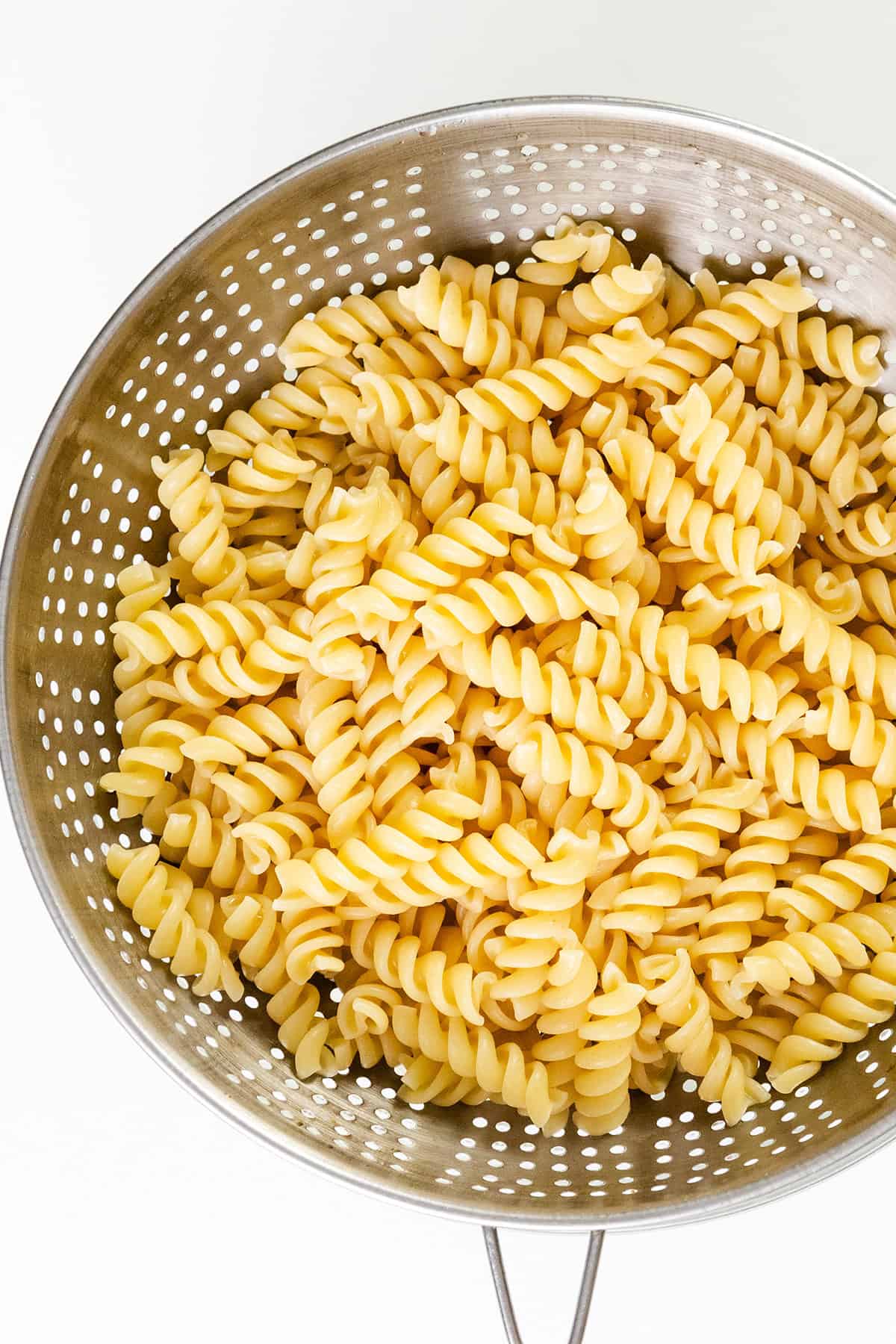 A metal colander filled with cooked rotini pasta, featuring tightly spiraled noodles, is placed on a white background. The pasta, perfect for a turkey casserole recipe, appears to be freshly drained and ready for mixing.