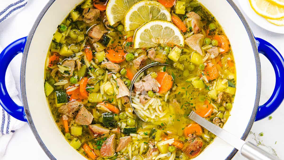 A pot of soup with chunks of meat, carrots, peas, zucchini, and orzo pasta, garnished with lemon slices. Perfect for comfort food recipes, this hearty meal sits in a pot with blue handles and a ladle resting inside.