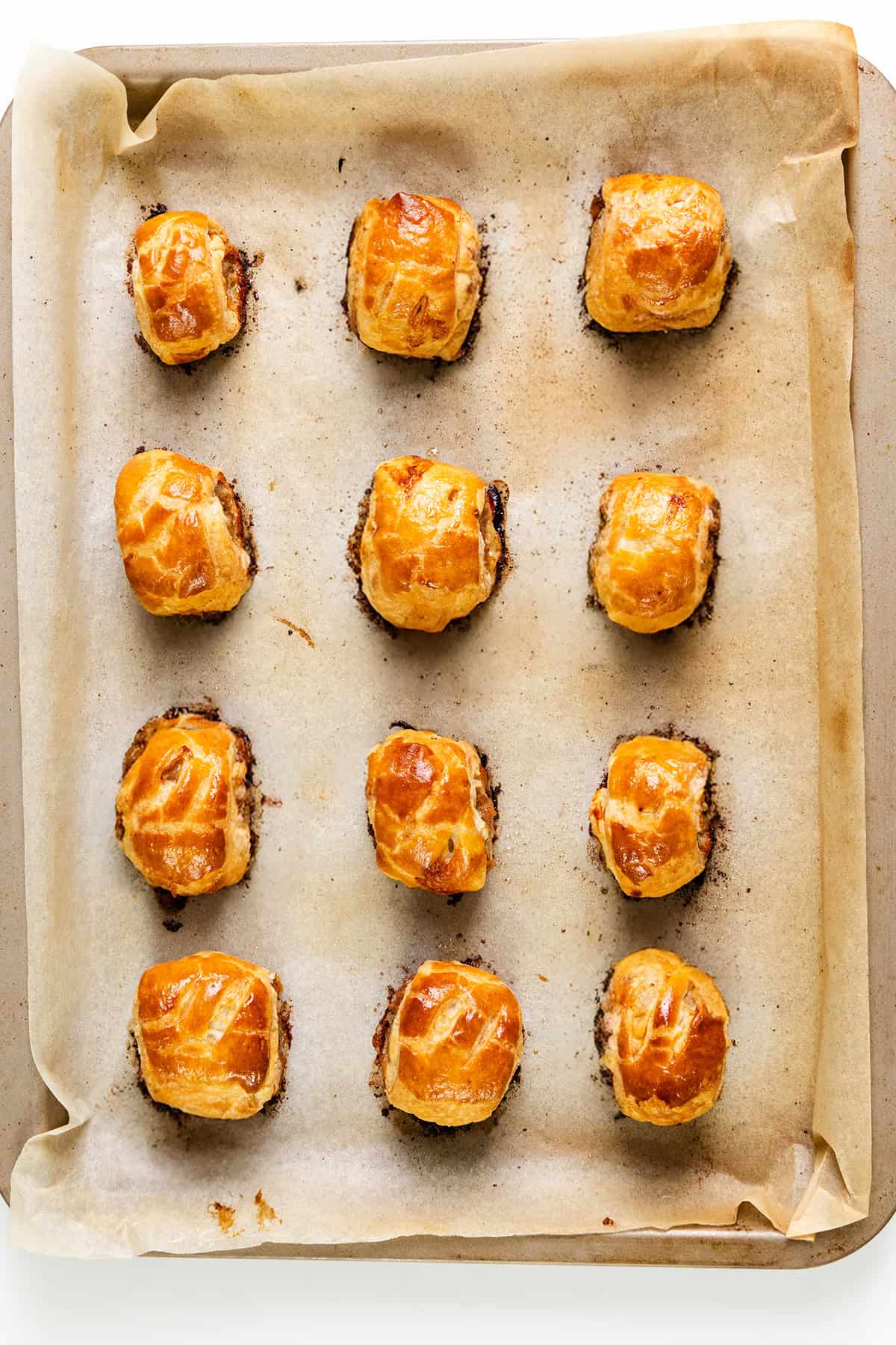 Twelve golden-brown homemade sausage rolls are arranged in a 4x3 grid on a parchment-lined baking tray. The rolls are evenly spaced and appear freshly baked, showcasing a glossy, flaky surface.
