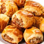 A plate filled with golden-brown homemade sausage rolls. The rolls are flaky and baked to a crisp, revealing a savory filling. They are arranged in a pile on a white plate, with a glossy sheen on the pastry.