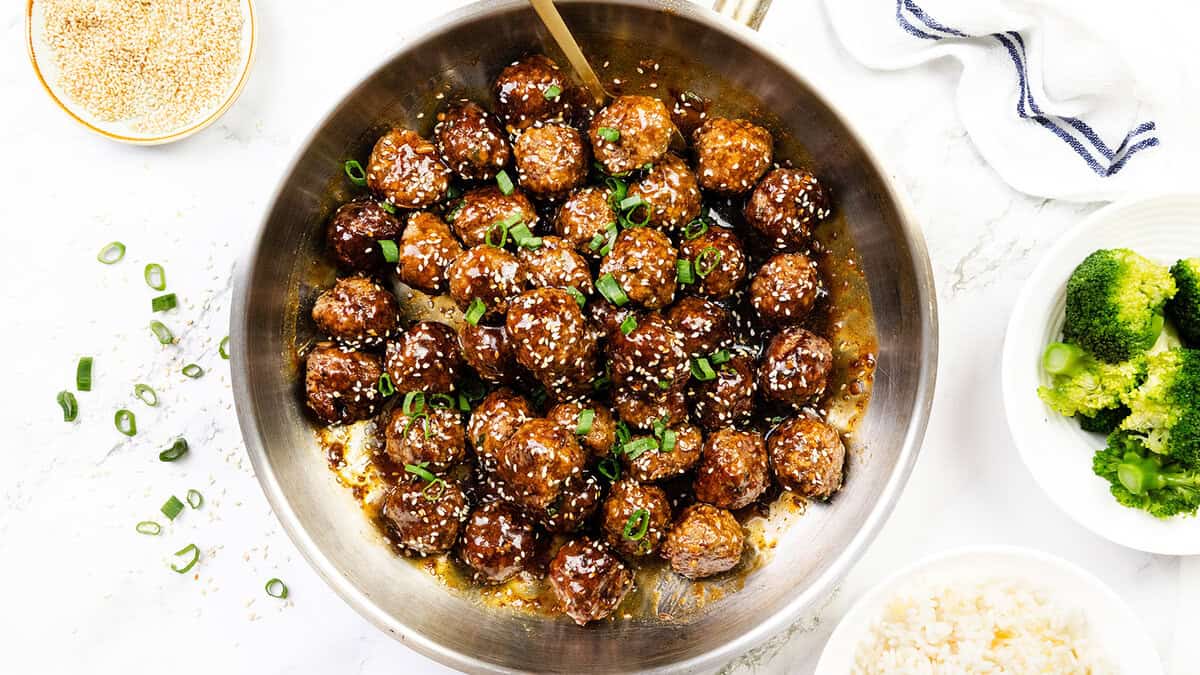 A skillet filled with glazed meatballs, garnished with sesame seeds and chopped green onions—perfect for fans of Asian recipes. Nearby, bowls of steamed broccoli, white rice, and extra sesame seeds sit on a white surface.