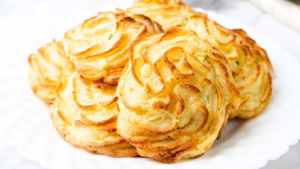 A plate of golden-brown duchess potatoes, delicately piped into swirled shapes. Each gourmet dish is crispy on the edges and fluffy inside, garnished with a hint of green herbs. The dish is set on a white textured plate.