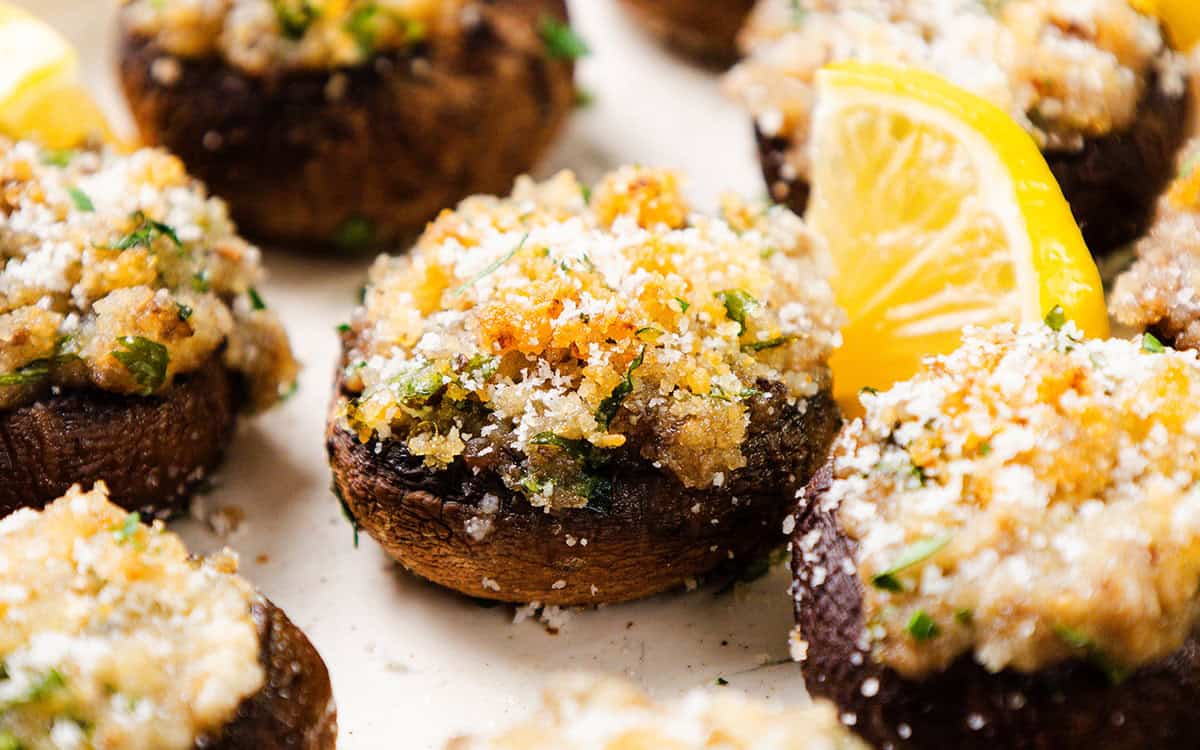 Close-up of stuffed mushrooms artfully arranged, their savory filling complemented by crispy breadcrumbs and herbs. Garnished with grated cheese and lemon wedges, these stuffed mushrooms lie on a pristine white surface, highlighting their delectable toppings.