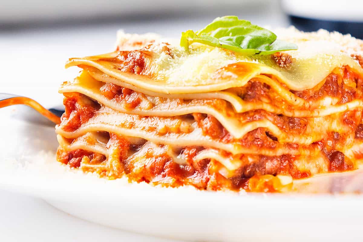 A close-up of a slice of Italian sausage lasagna on a white plate. The cheesy delight boasts multiple layers of pasta, rich meat sauce, and melted cheese, topped with a sprig of fresh basil and garnished with a sprinkle of grated cheese.