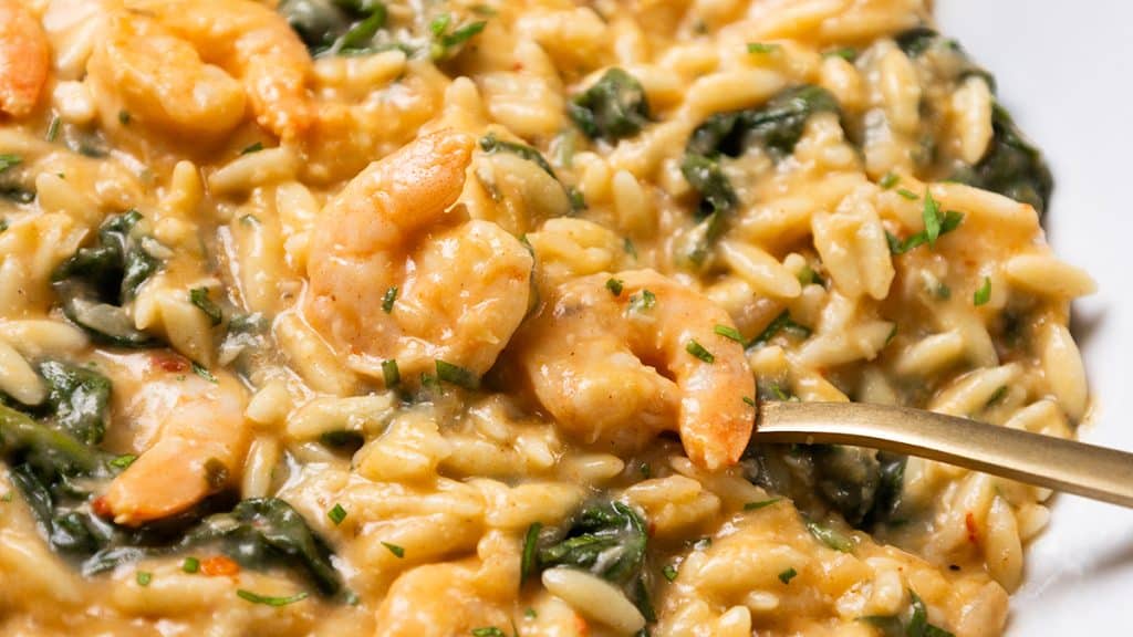 Creamy orzo pasta mixed with shrimp and spinach, garnished with chopped herbs. Perfect for Quick Prep meals, this dish boasts a rich, cheesy texture. A gold fork is partially visible, ready to take a bite.