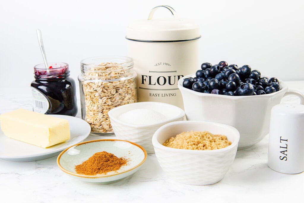 A variety of baking ingredients for a new recipe are arranged on a white surface, including blueberries, oats, flour, butter, jam, sugar, brown sugar, cinnamon, and a salt shaker.