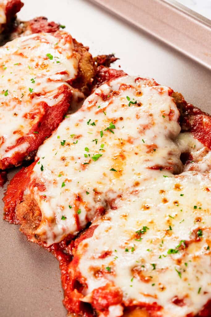 Close-up of classic chicken parmesan on a baking tray. The dish features crispy breaded chicken cutlets topped with marinara sauce and melted mozzarella cheese, sprinkled with chopped parsley. The cheese is golden and slightly browned.