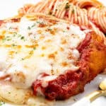 A classic chicken parmesan dish, topped with melted cheese and rich tomato sauce, is garnished with herbs, sitting beside a serving of spaghetti. A gold fork lies ready for the feast.