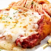 A classic chicken parmesan dish, topped with melted cheese and rich tomato sauce, is garnished with herbs, sitting beside a serving of spaghetti. A gold fork lies ready for the feast.