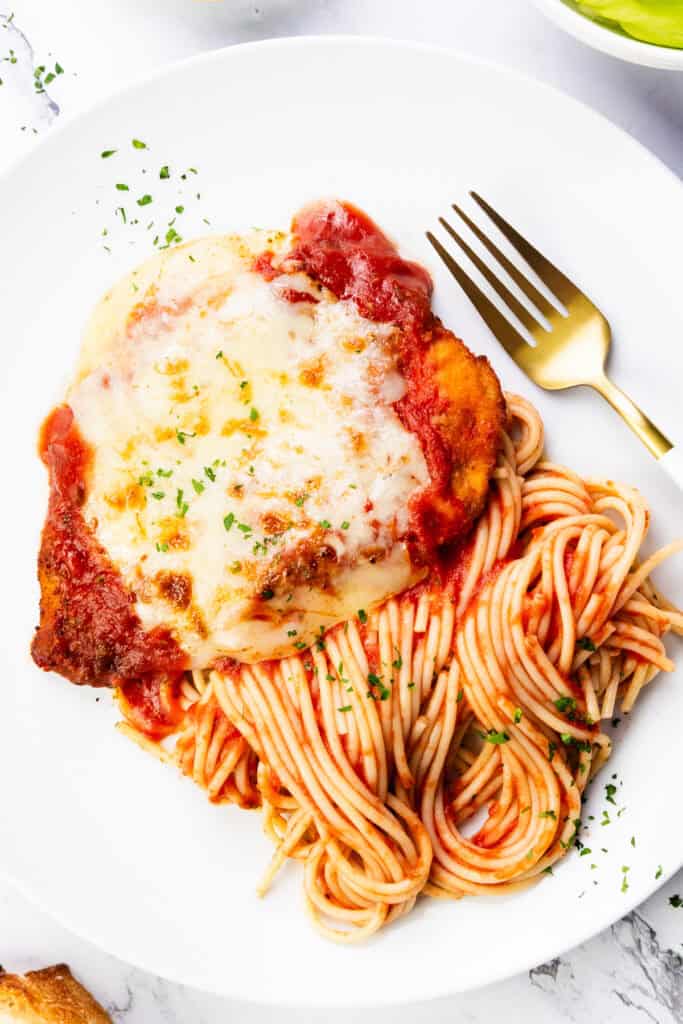 A plate of Classic Chicken Parmesan features spaghetti with marinara sauce and a piece of chicken topped with melted cheese. A fork rests beside the chicken, and the dish is garnished with chopped herbs. A slice of bread peeks out from the corner.