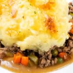 Close-up of a plate of cottage pie, showcasing a golden-brown mashed potato topping over a savory filling of ground meat, peas, carrots, and rich gravy. The dish appears hot and freshly baked.