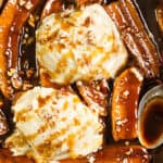 Scoops of vanilla ice cream topped with caramel sauce, surrounded by caramelized banana slices in a delightful Bananas Foster style, and sprinkled with chopped nuts. A spoon rests in the rich, saucy mixture.