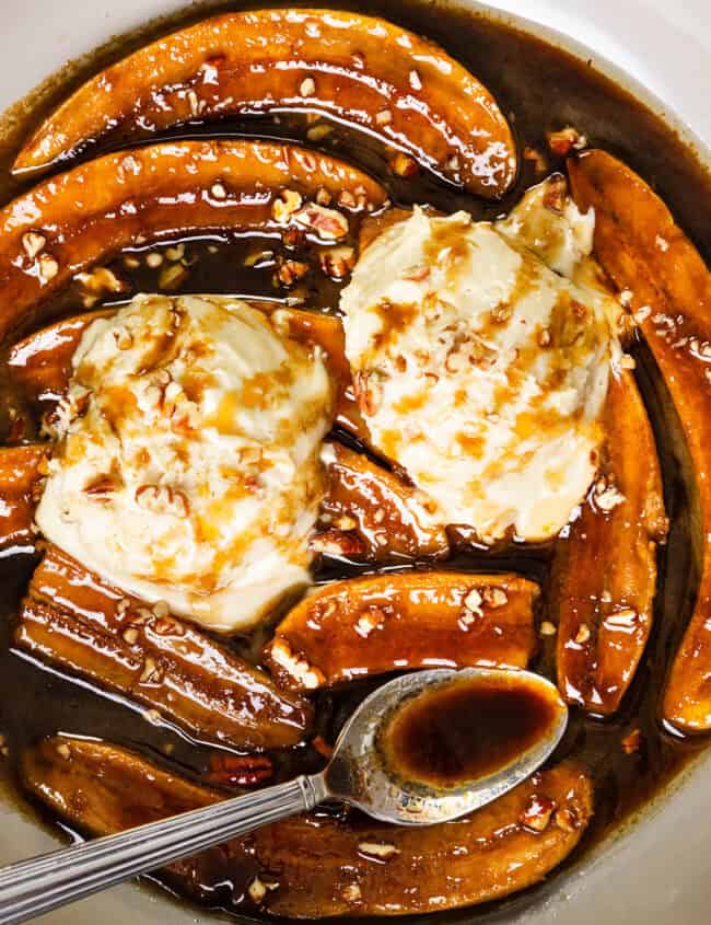 A plate of Bananas Foster showcases caramelized banana slices in a rich brown sauce, crowned with scoops of vanilla ice cream and a sprinkle of chopped nuts. A spoon rests in the sauce, capturing the dessert's indulgent texture and sweetness.