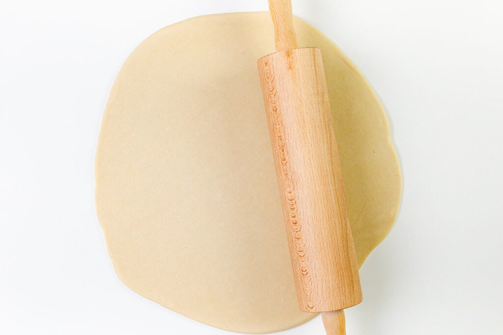 A wooden rolling pin rolling out a sheet of dough for a buttery crust on a white surface, perfect for making cheese quiche.
