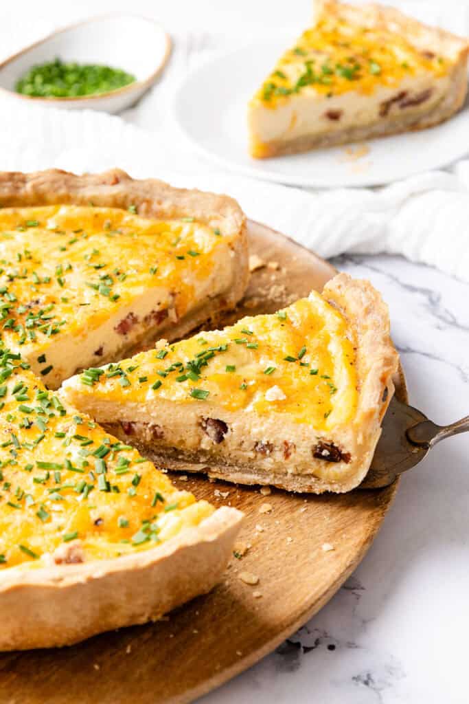 A slice of Cheese Quiche with a golden, cheesy top and visible bacon pieces is being lifted from a wooden serving board. The quiche, featuring a buttery crust and garnished with chopped chives, sits beside another slice in the background.