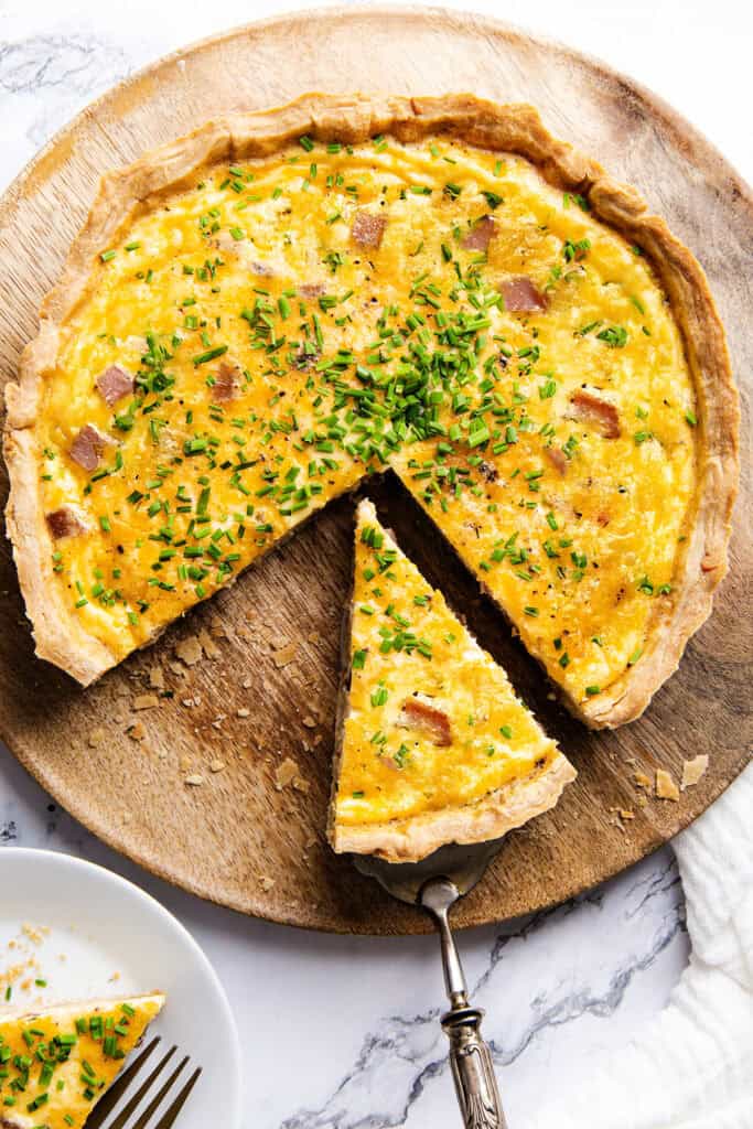 A cheese quiche with a buttery crust, filled with eggs, ham, and herbs, sits on a wooden board. One slice is cut and served, garnished with chopped green chives. A plate with a slice and a fork is nearby.