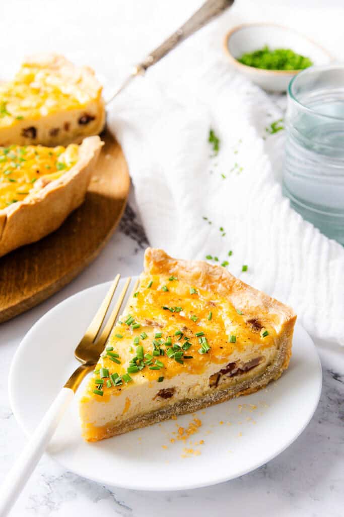 A slice of cheese quiche with herbs on top sits on a white plate with a fork, next to a glass of water, showcasing its buttery crust, with the rest of the quiche and a bowl of chopped herbs in the background.