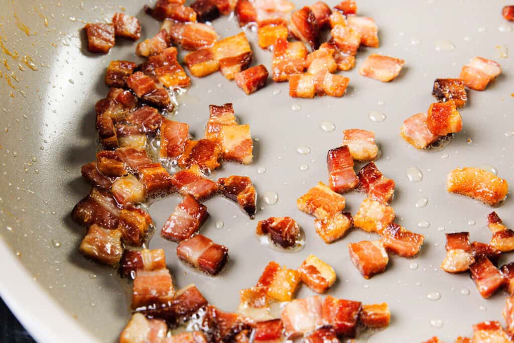 Small, crispy bacon pieces are sizzling in a frying pan, with visible oil and fat rendering around them on the pan's surface—perfect for topping a cheese quiche or adding crunch to a buttery crust.