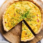 A Cheese Quiche with a buttery crust and yellow filling, topped with chopped chives, sits on a wooden board. One slice is cut and slightly pulled out from the whole quiche.