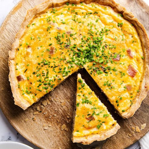 A Cheese Quiche with a buttery crust and yellow filling, topped with chopped chives, sits on a wooden board. One slice is cut and slightly pulled out from the whole quiche.