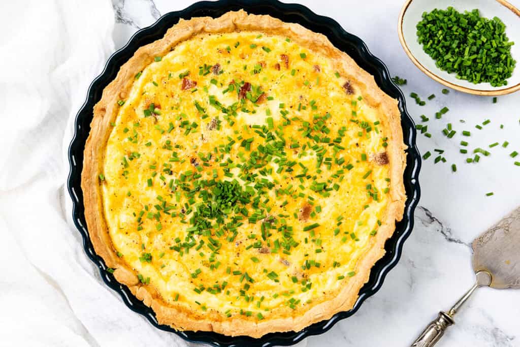 A cheese quiche with a buttery crust sits in a round black dish, topped with chopped chives, on a marble surface next to a bowl of extra chives and a metal pie server.