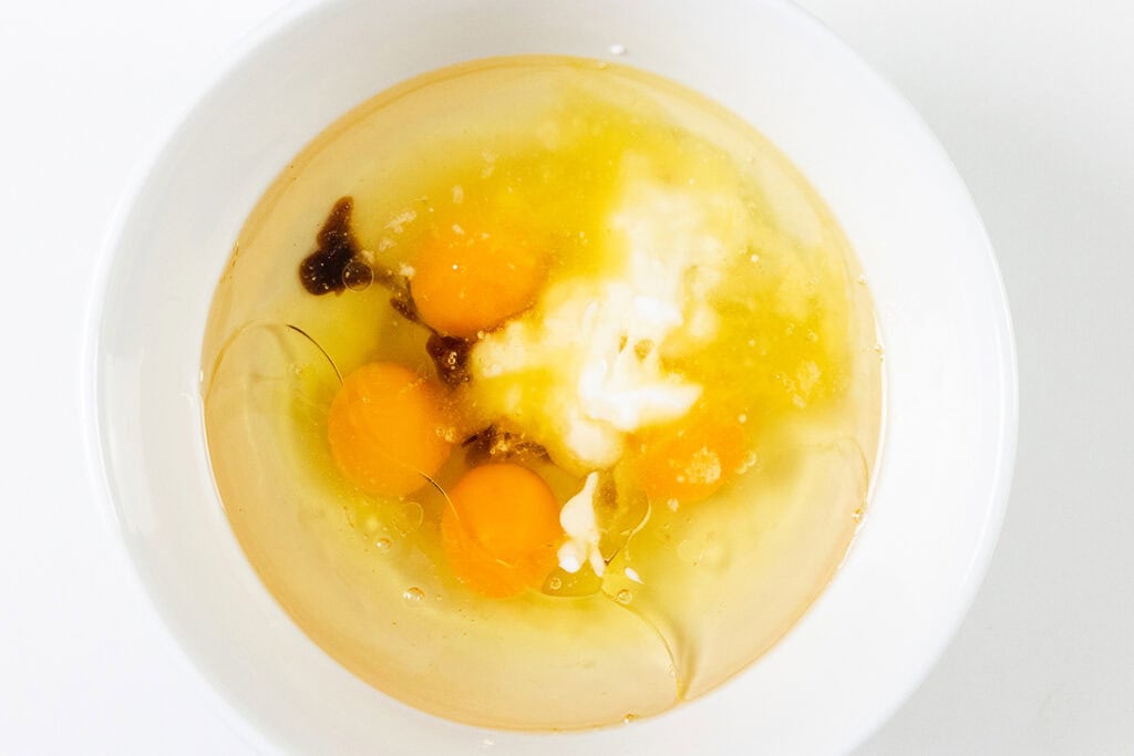 A white bowl containing cracked eggs with yolks and whites, clear liquid, and dark specks, likely vanilla extract, sits on a white surface—perfect beginnings for a decadent carrot cake or cream cheese frosting.