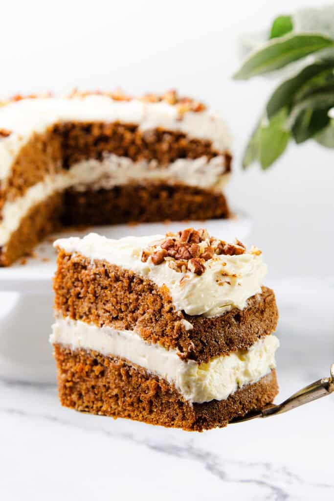 A slice of carrot cake topped with luscious cream cheese frosting is being lifted by a cake server. The two-layer delight, filled and crowned with creamy frosting and chopped nuts, tantalizes in the foreground, while the rest of this carrot cake masterpiece sits on a white cake stand.