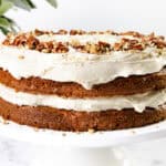 A two-layer carrot cake with cream cheese frosting and a sprinkle of chopped pecans rests elegantly on a white cake stand, while a leafy plant adds a touch of greenery in the background.