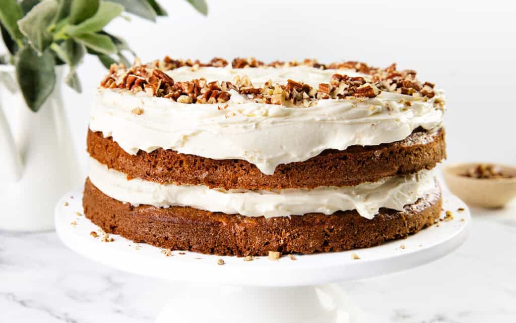 Two-layered carrot cake with rich cream cheese frosting, topped with chopped pecans, displayed on a white cake stand. A plant and a bowl of pecans are in the background.