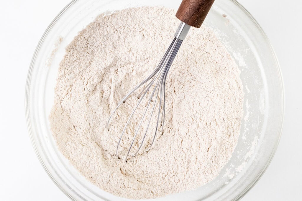A clear glass bowl containing a mound of flour, part of a delicious carrot cake recipe, is seen from above. A metal whisk with a wooden handle is partially submerged in the flour, hinting at mixing in progress. The background is a plain white surface, ready for the magic of cream cheese frosting to follow.