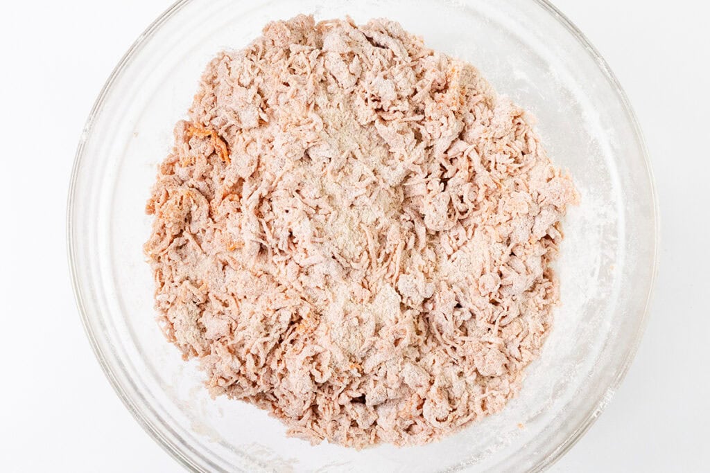 A glass bowl containing a mixture of shredded raw chicken, dusted with flour, sits on a white surface. The flour lightly coats the chicken, reminiscent of how cream cheese frosting wraps around every layer of a carrot cake.