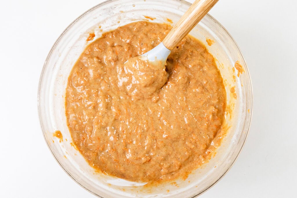 A glass bowl brimming with light brown carrot cake batter, where a wooden-handled spatula rests in the thick mixture, speckled with tiny pieces of carrot. The bowl sits invitingly on a white surface, just waiting to be topped with luscious cream cheese frosting.