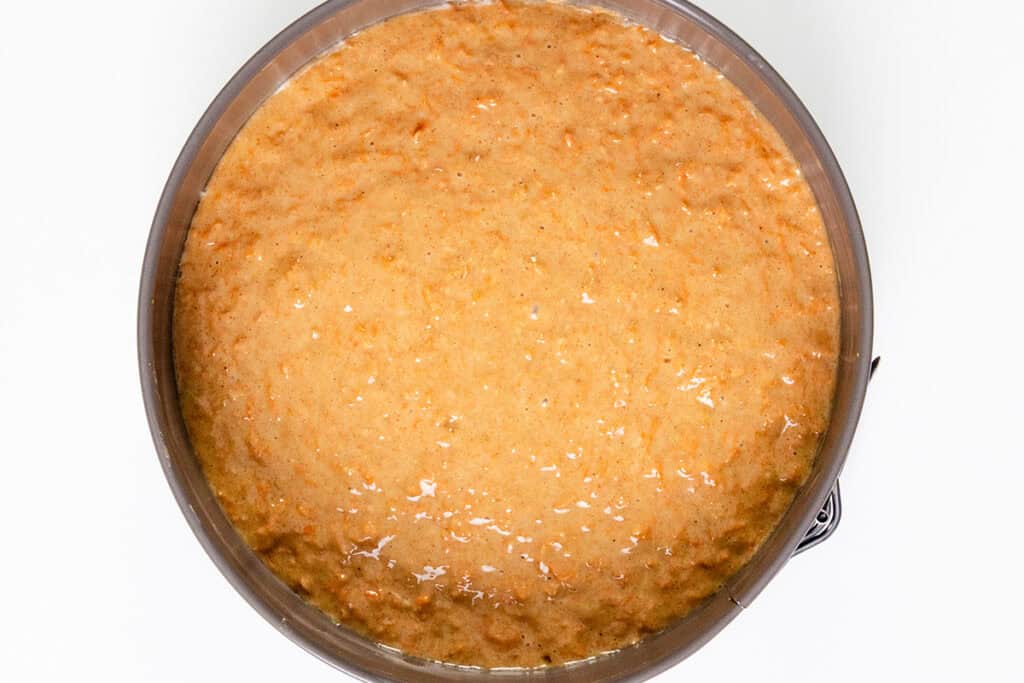 A round baking pan filled with smooth, light brown carrot cake batter, ready to be baked. The batter appears moist and slightly textured, promising a delicious treat once topped with luscious cream cheese frosting.