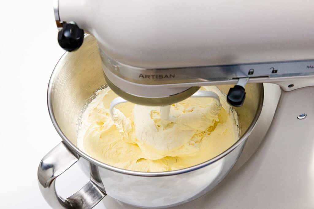 A stand mixer with a metal bowl is shown mixing smooth, creamy yellow cake batter, ready to become a delicious carrot cake. The word "ARTISAN" is visible on the mixer. The batter appears light and fluffy, perfect for pairing with rich cream cheese frosting against a plain white background.
