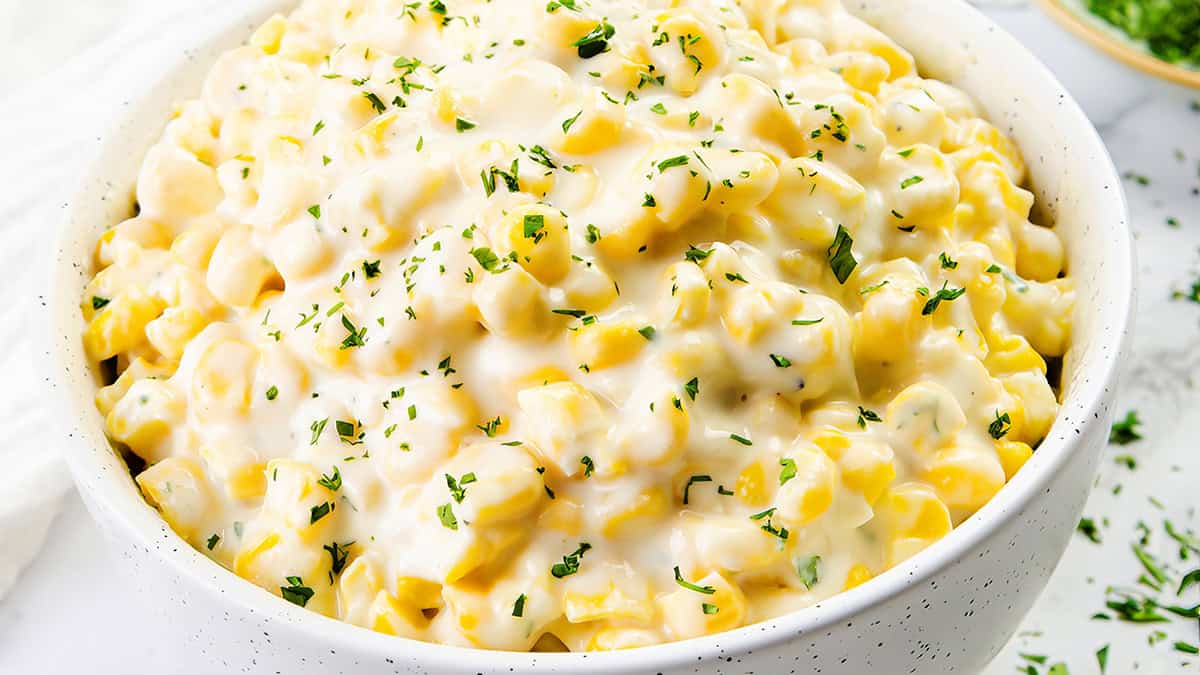 A white bowl filled with creamy, cheesy corn topped with chopped green herbs, set on a light surface.