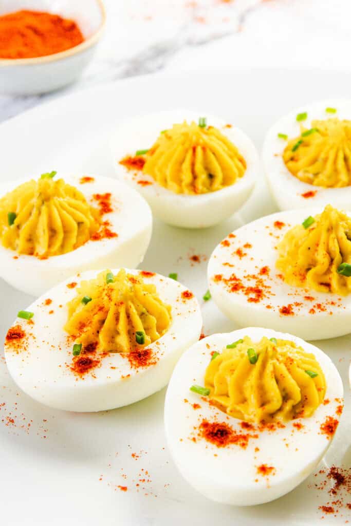 A plate of Southern deviled eggs is beautifully garnished with paprika and chives on a white dish. The eggs are halved with creamy yellow filling piped neatly on top. A small bowl of paprika, a staple in Southern recipes, is visible in the background.