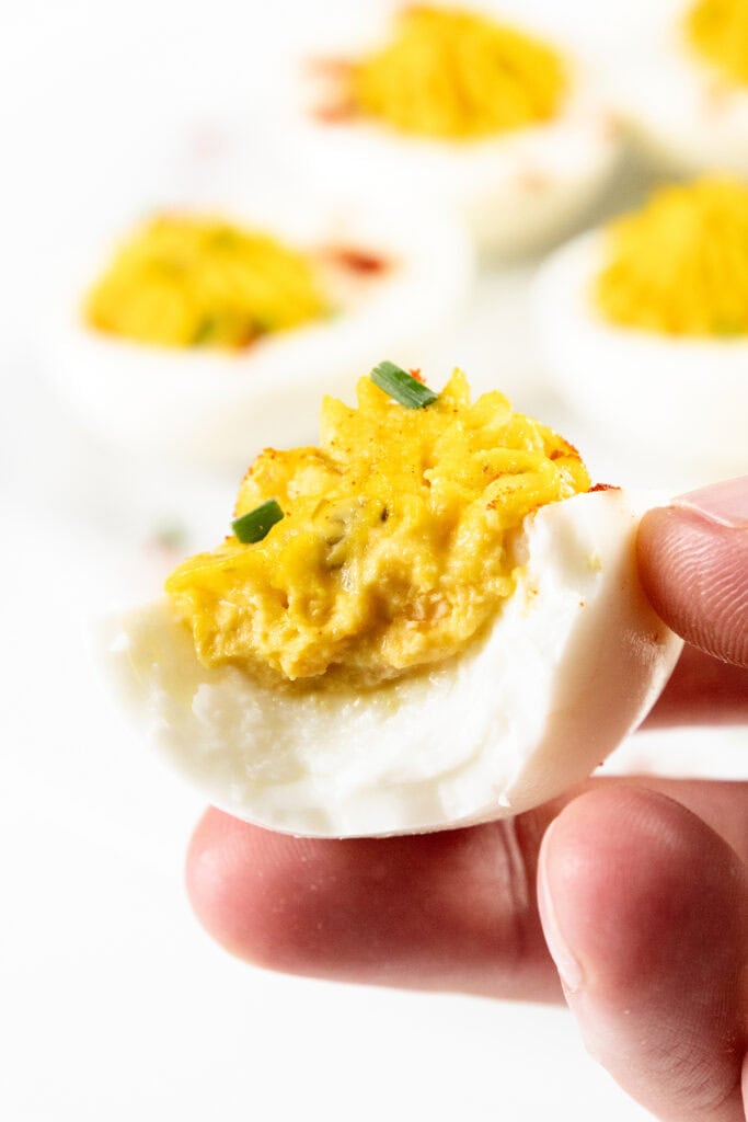 Close-up of a hand holding half of a classic Southern deviled egg, topped with a creamy yellow filling and garnished with a small green herb piece. More deviled eggs, perfect for any recipe collection, are blurred in the background.