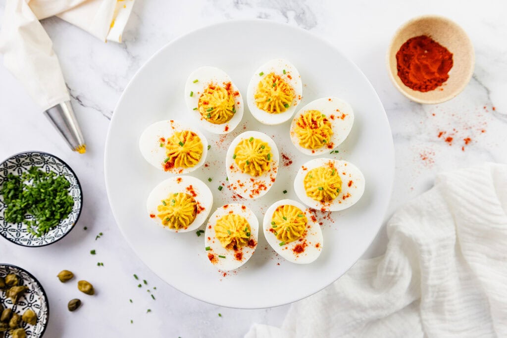 A plate of Southern Deviled Eggs garnished with paprika and chives sits elegantly on a white marble surface. Nearby, small bowls hold red spice, chopped herbs, and capers, complemented by a piping bag and crisp white cloth.