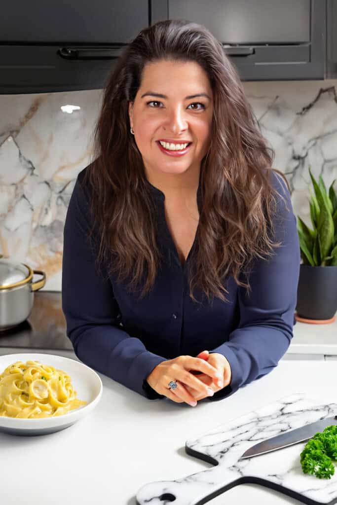 Erren Hart with long hair, smiling, leans on a kitchen counter. In front, there's a bowl of pasta. The background, embodying our philosophy of elegance meets function, features marble-patterned walls, a plant, and a cutting board with greens in this modern kitchen setting.