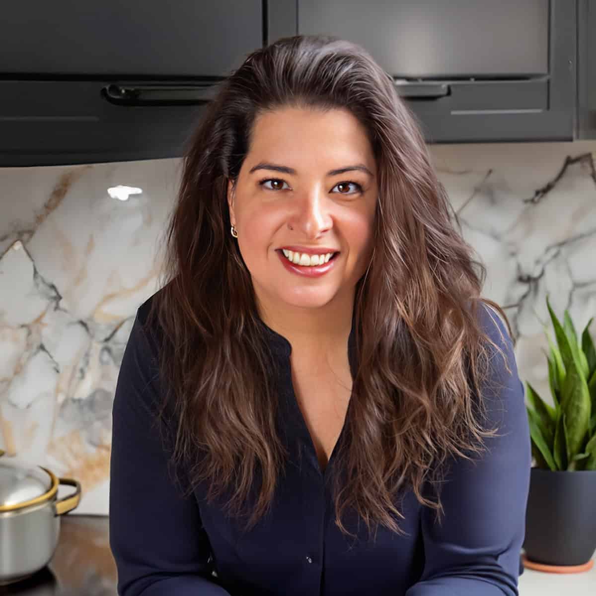 Erren Hart with long hair, smiling, leans on a kitchen counter. In front, there's a bowl of pasta. The background, embodying our philosophy of elegance meets function, features marble-patterned walls, a plant, and a cutting board with greens in this modern kitchen setting.