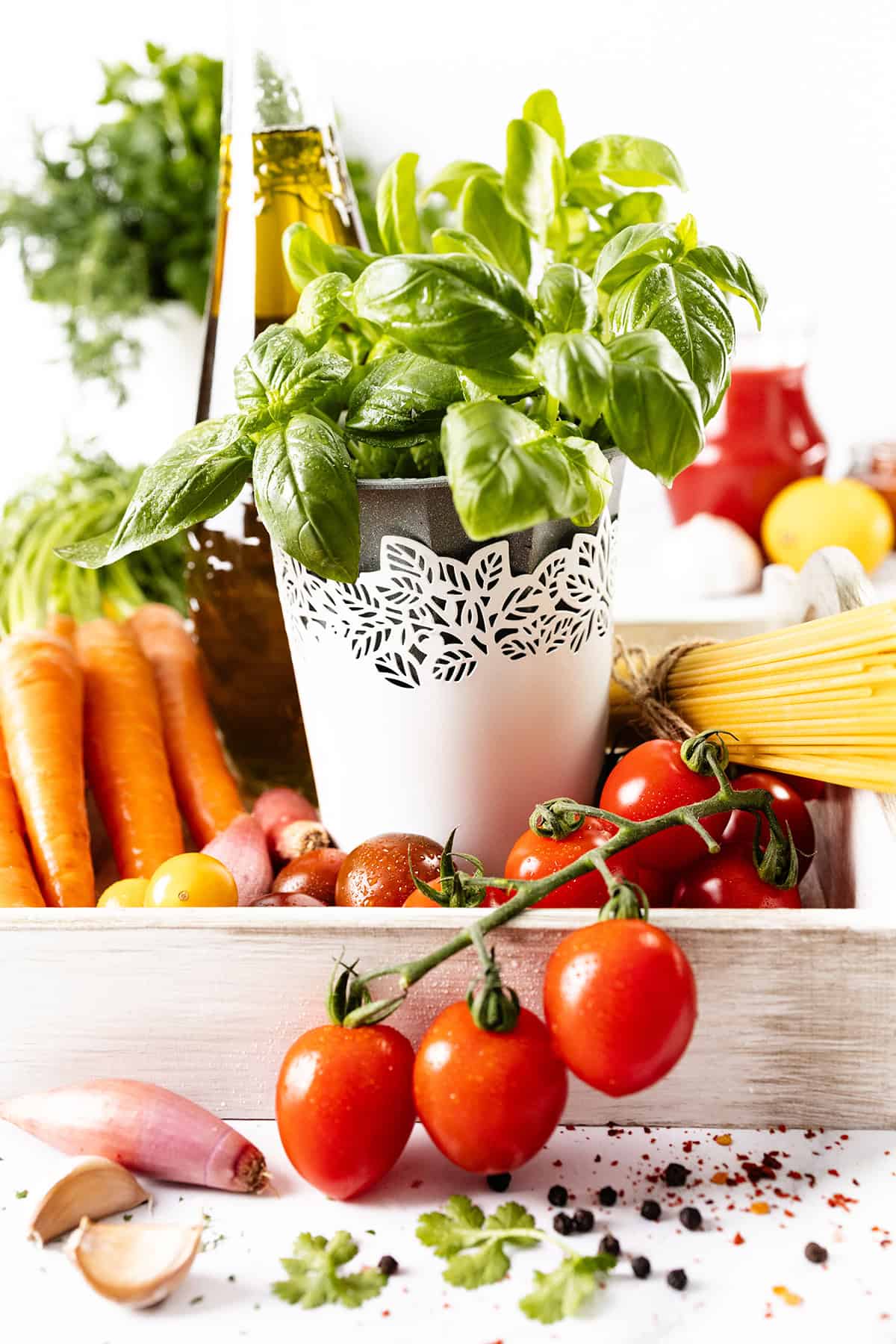 An arrangement of fresh vegetables and herbs: a pot of basil, cherry tomatoes, carrots, and garlic. A bottle of olive oil and a bundle of uncooked spaghetti are in the background, with peppercorns scattered around.